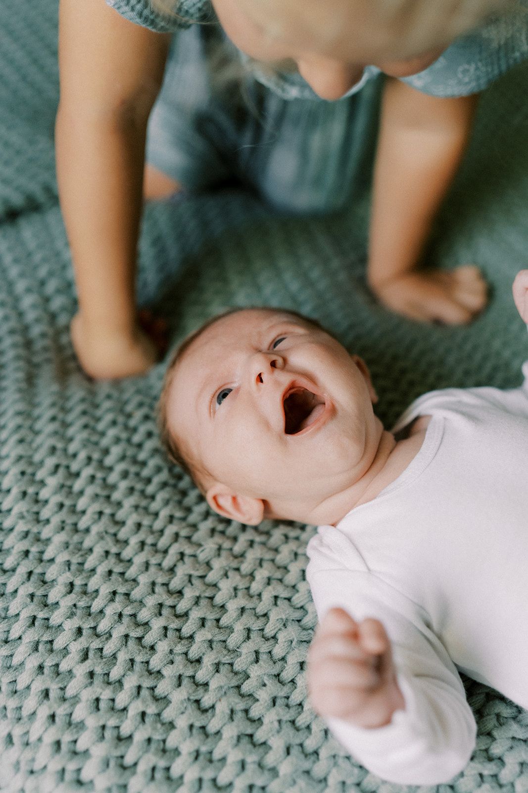 Laughing newborn baby in white onesie lying on teal knit blanket with older sibling nearby.