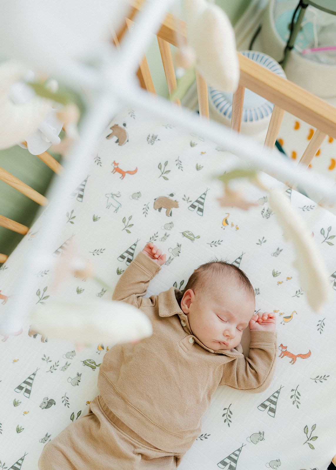 Newborn baby sleeping peacefully in a woodland-themed crib with a forest animals sheet and hanging mobile above.