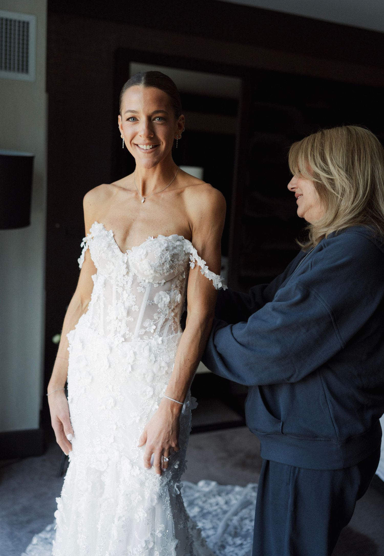 Bride smiling in floral off-shoulder wedding gown being assisted by a woman in a navy blazer.