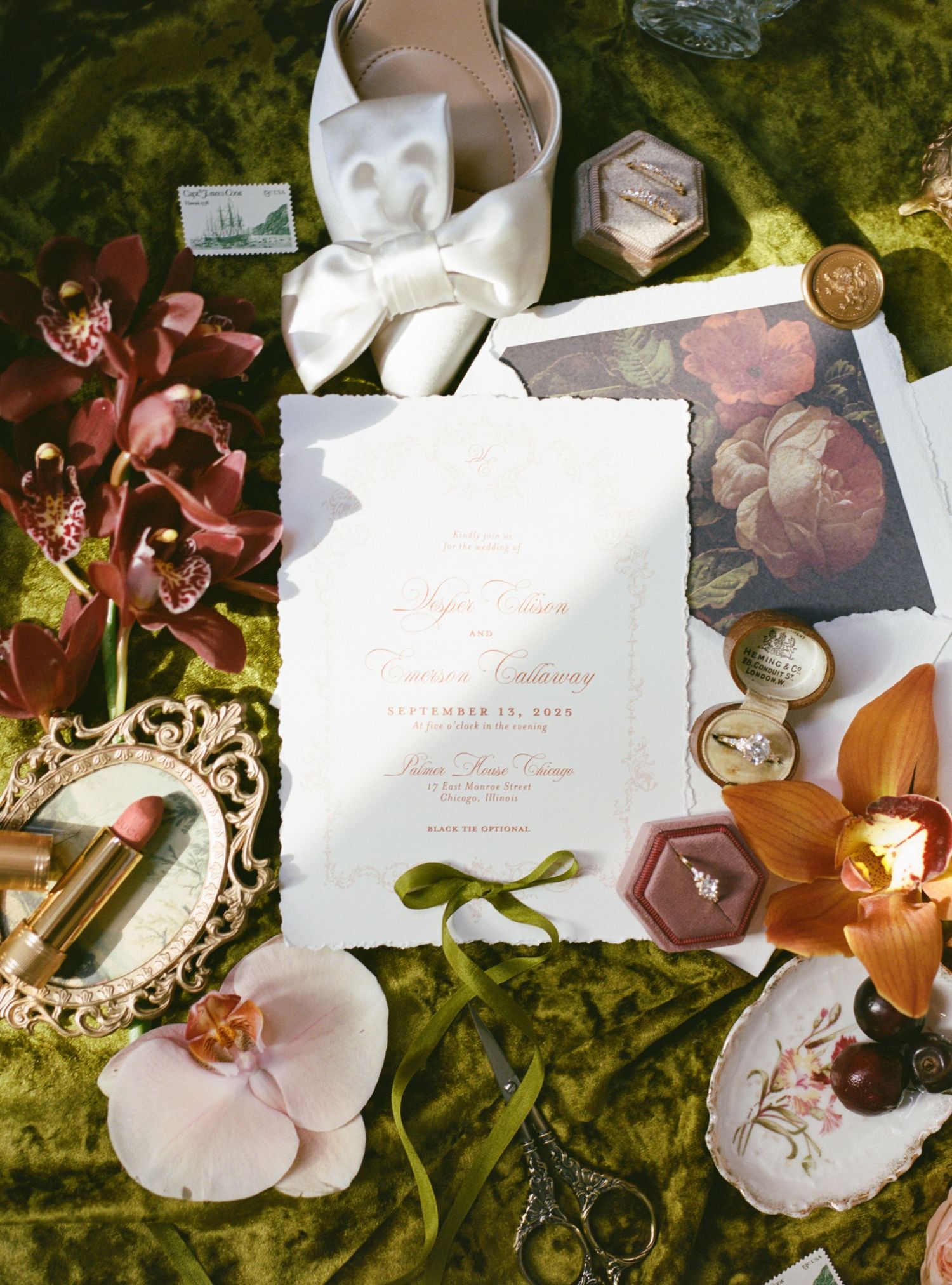 Elegant wedding invitation suite on green velvet with orchids, rings, lipstick, and vintage accessories.
