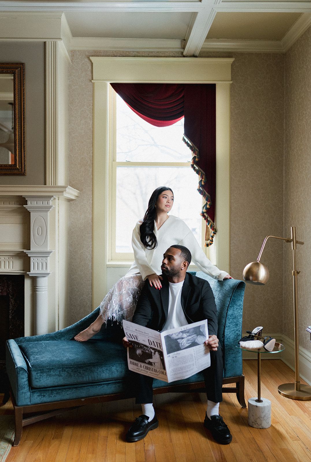 Elegant couple posing in a vintage room, bride by window and groom reading newspaper on teal chaise.