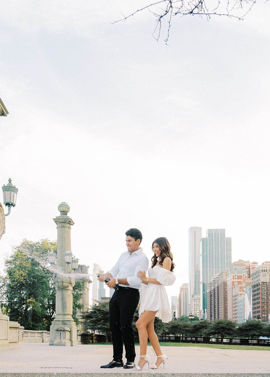 Couple in white outfits pop champagne bottle outdoors with Chicago city skyline in background.