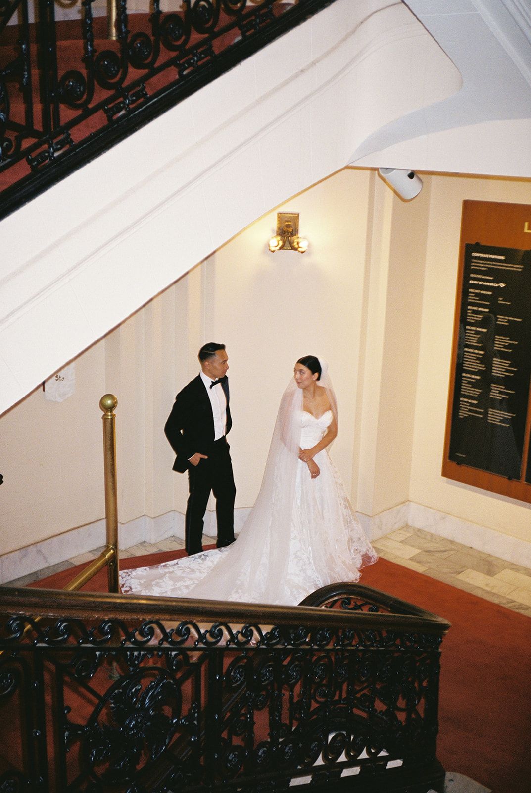 Bride and groom share a romantic moment on grand staircase, she in lace gown with veil, he in black tuxedo.