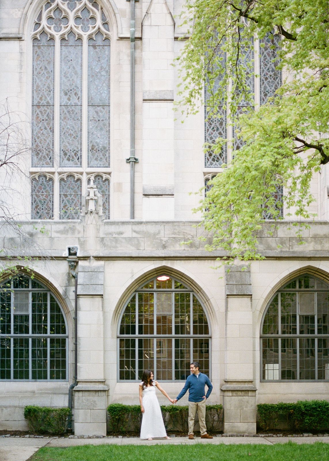Couple holding hands outside a Gothic-style stone church with arched windows.