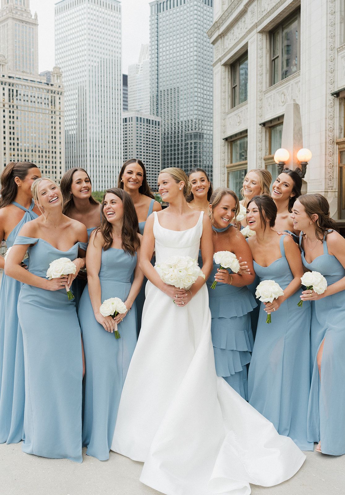 Bride in white gown poses with bridesmaids in dusty blue dresses against Chicago city skyline backdrop.