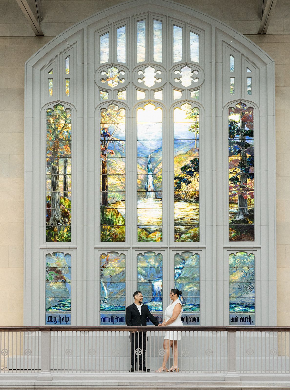 Couple stands before a stunning stained glass window in an elegant church during their wedding engagement session.