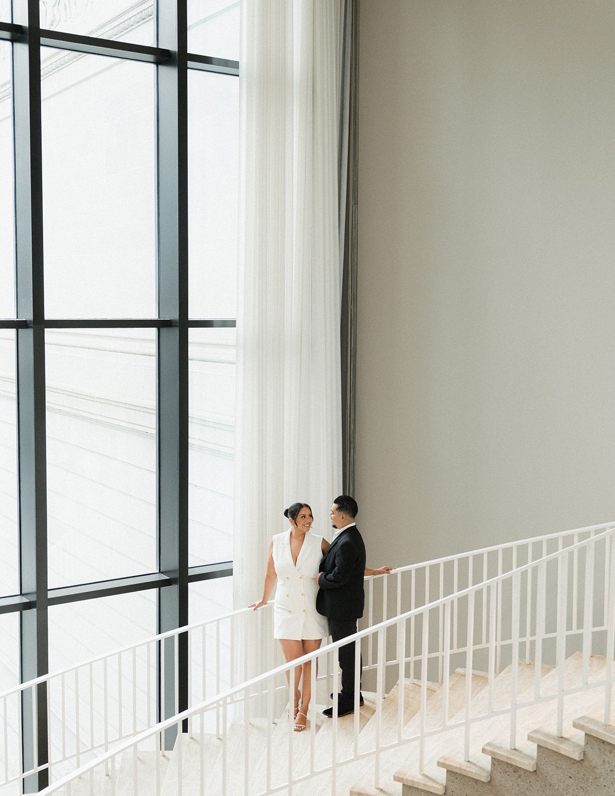 Elegant couple in wedding attire on a grand staircase with floor-to-ceiling windows.