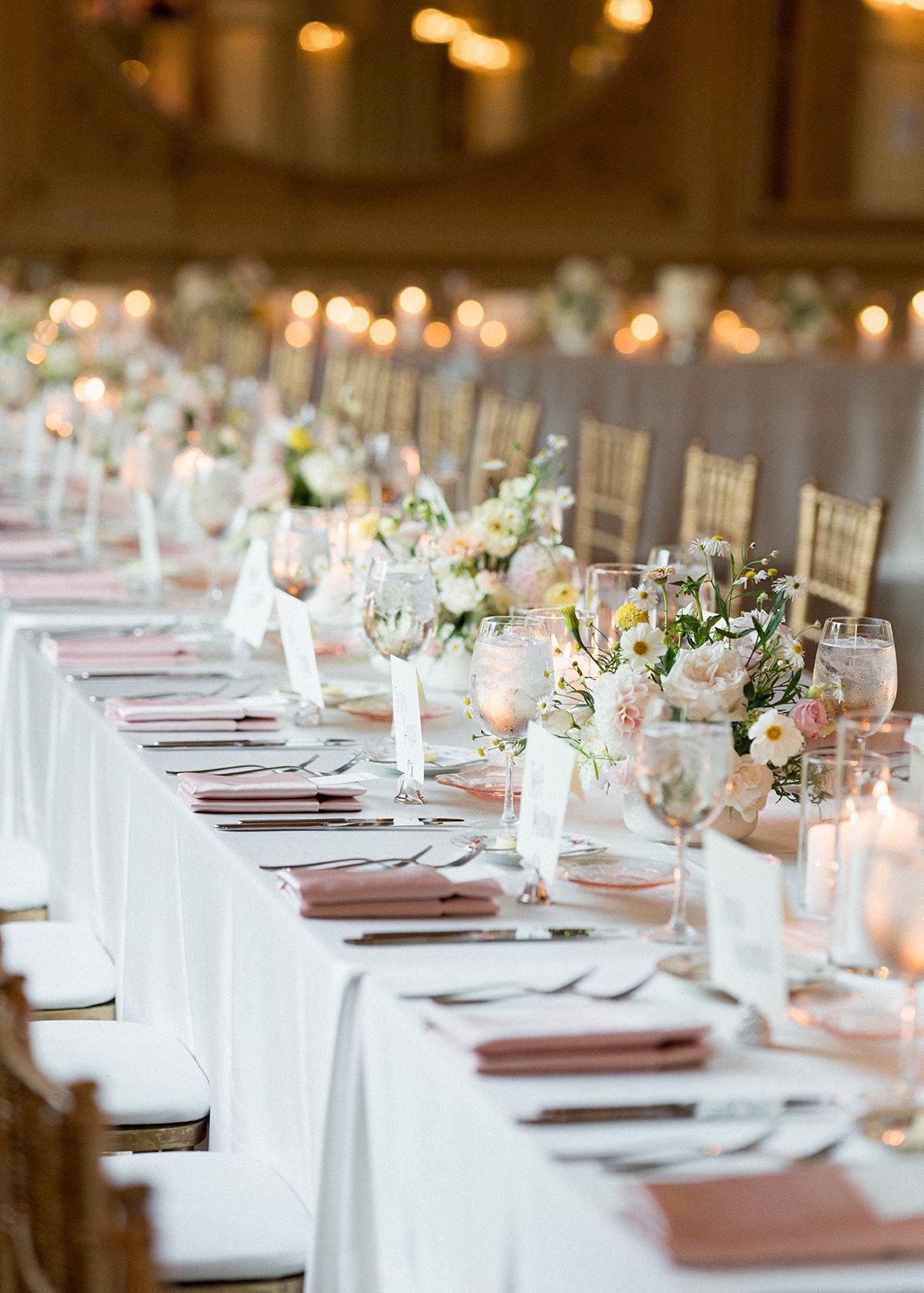 Elegant wedding reception table with white linens, blush napkins, floral centerpieces, crystal glassware, and candlelight.