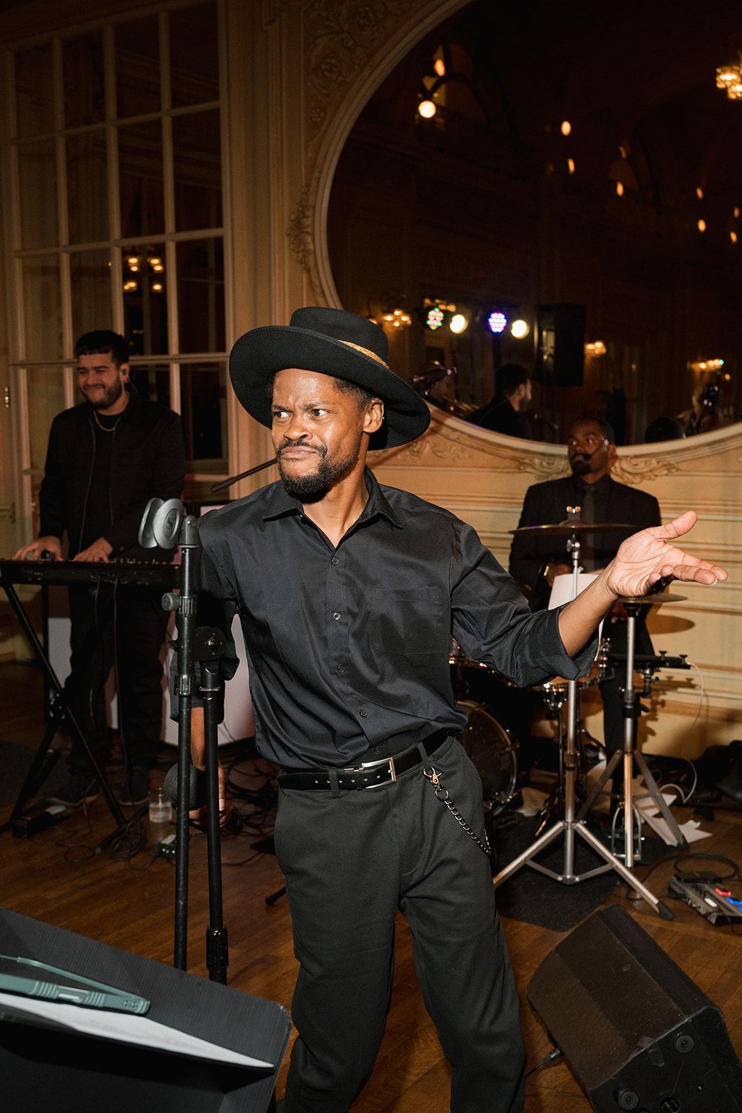 A man in black outfit and cowboy hat performs at microphone with band playing keyboard and drums behind him.