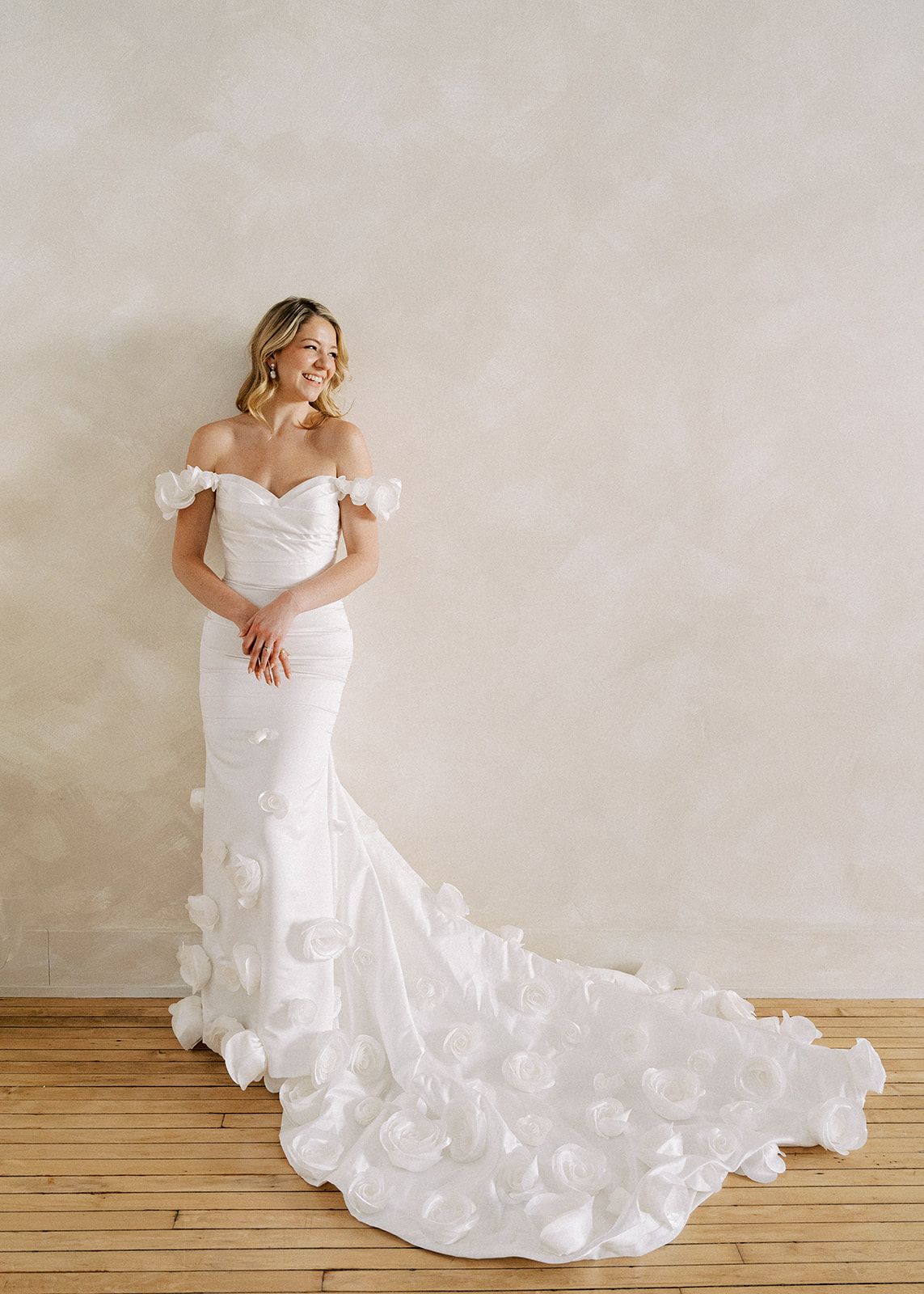 Elegant bride in off-shoulder mermaid wedding gown with floral petal train posing in a bright studio.