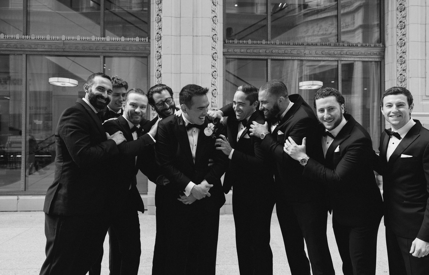 Black and white photo of groom with groomsmen in tuxedos laughing together outside elegant building.