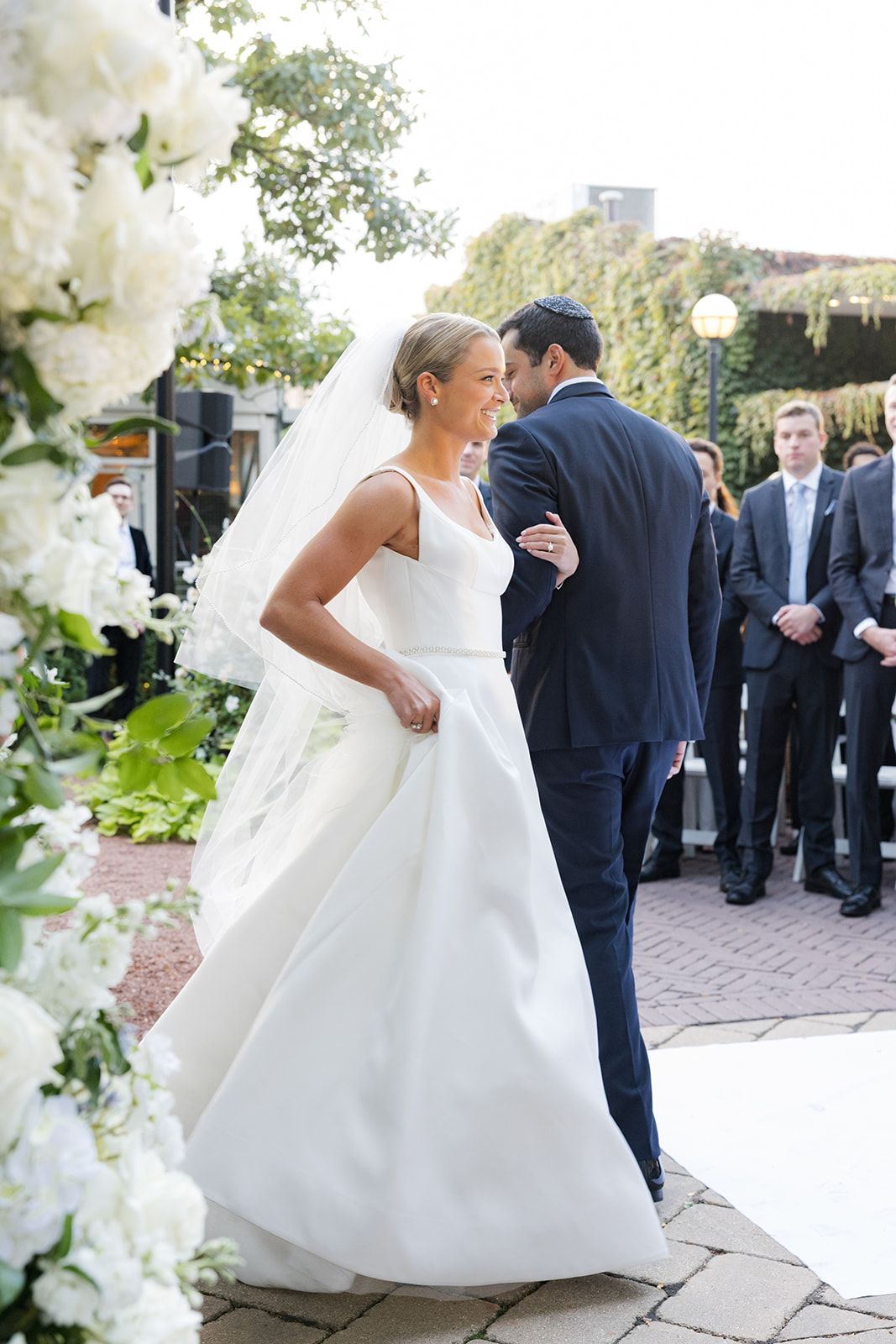 Bride and groom share intimate moment at outdoor garden ceremony surrounded by lush white floral arrangements.