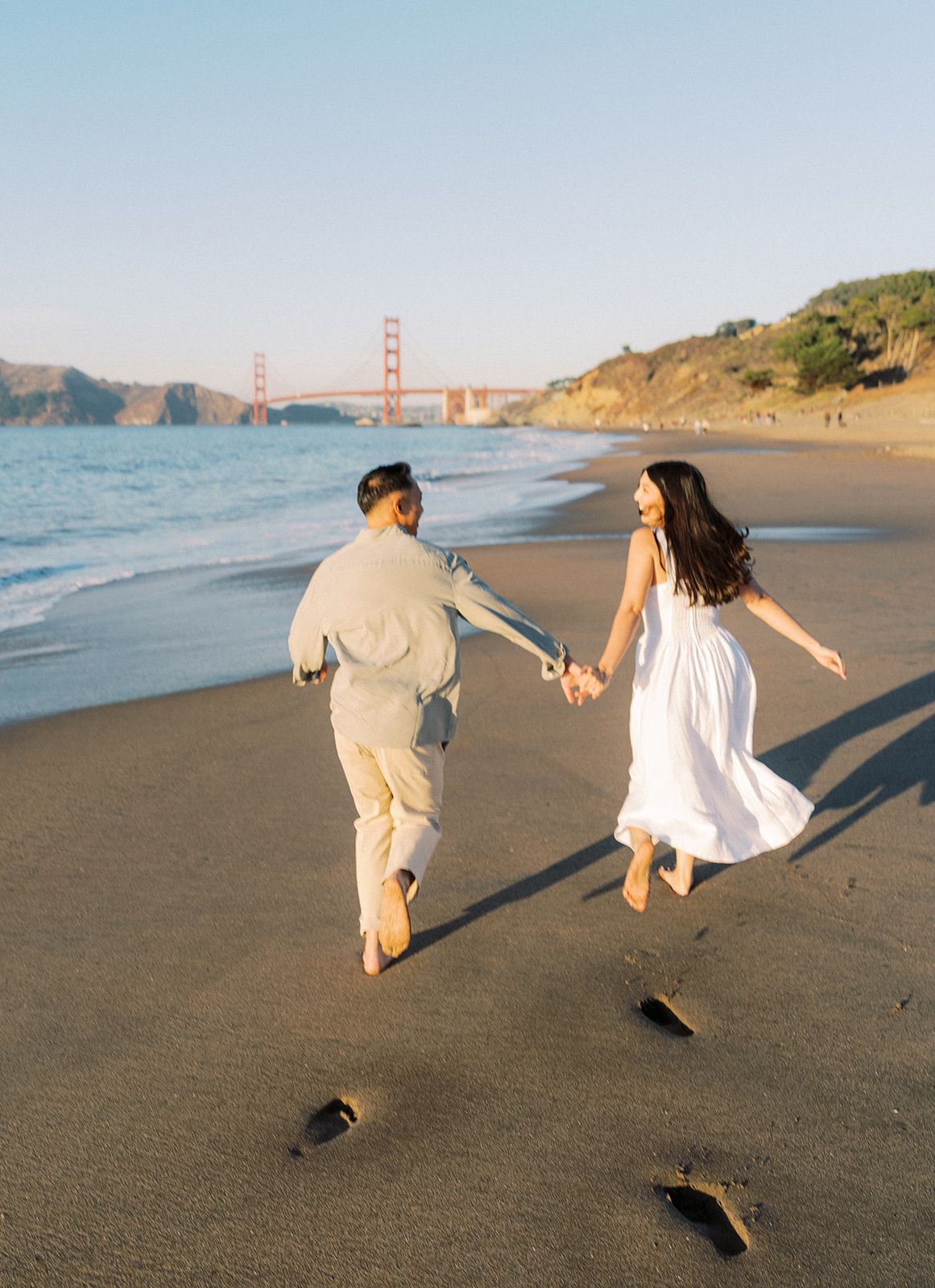 Couple holding hands running on a beach with the Golden Gate Bridge in the background.