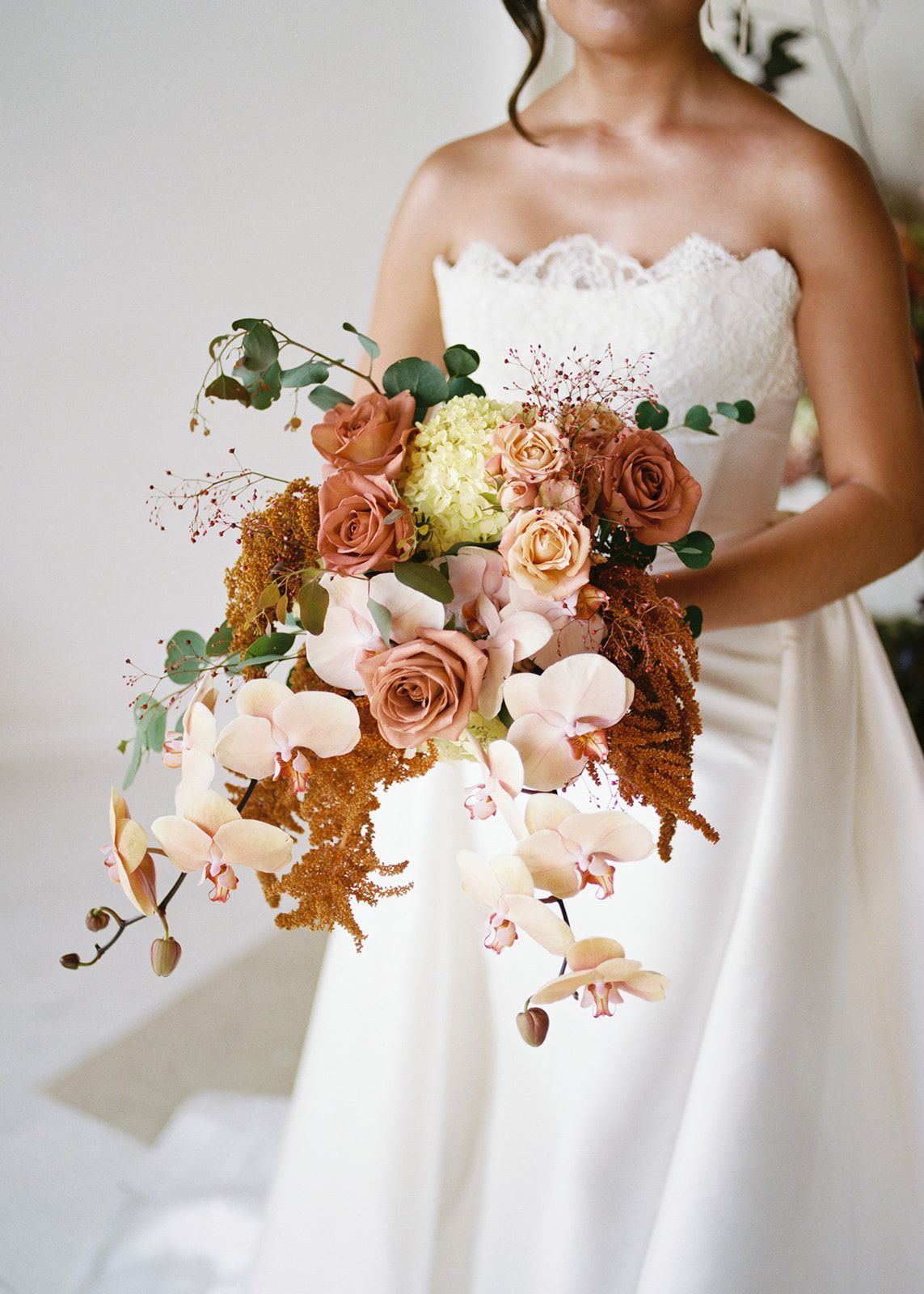 Bride holding cascading wedding bouquet with terracotta roses, orchids, and eucalyptus in lace strapless gown.