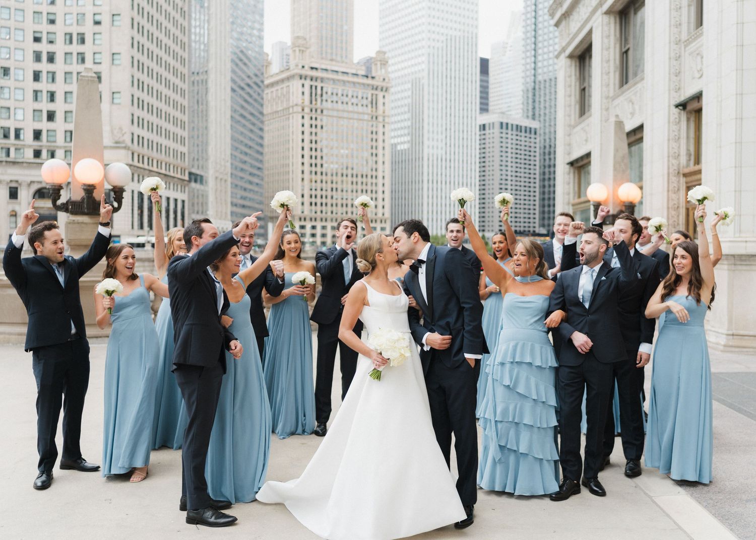 Wedding party cheers with bouquets raised as bride and groom kiss on Chicago Riverwalk terrace.