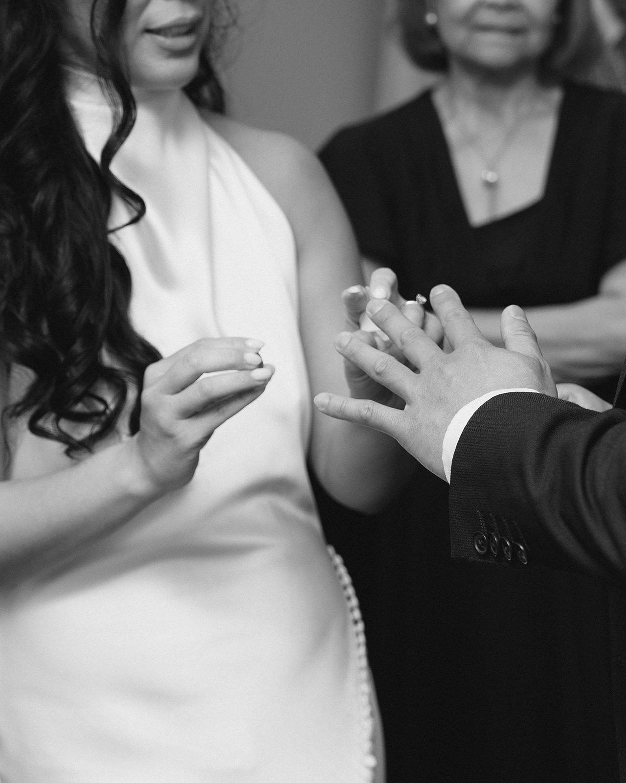 Black and white photo of a bride placing a wedding ring on the groom's finger during a ceremony.