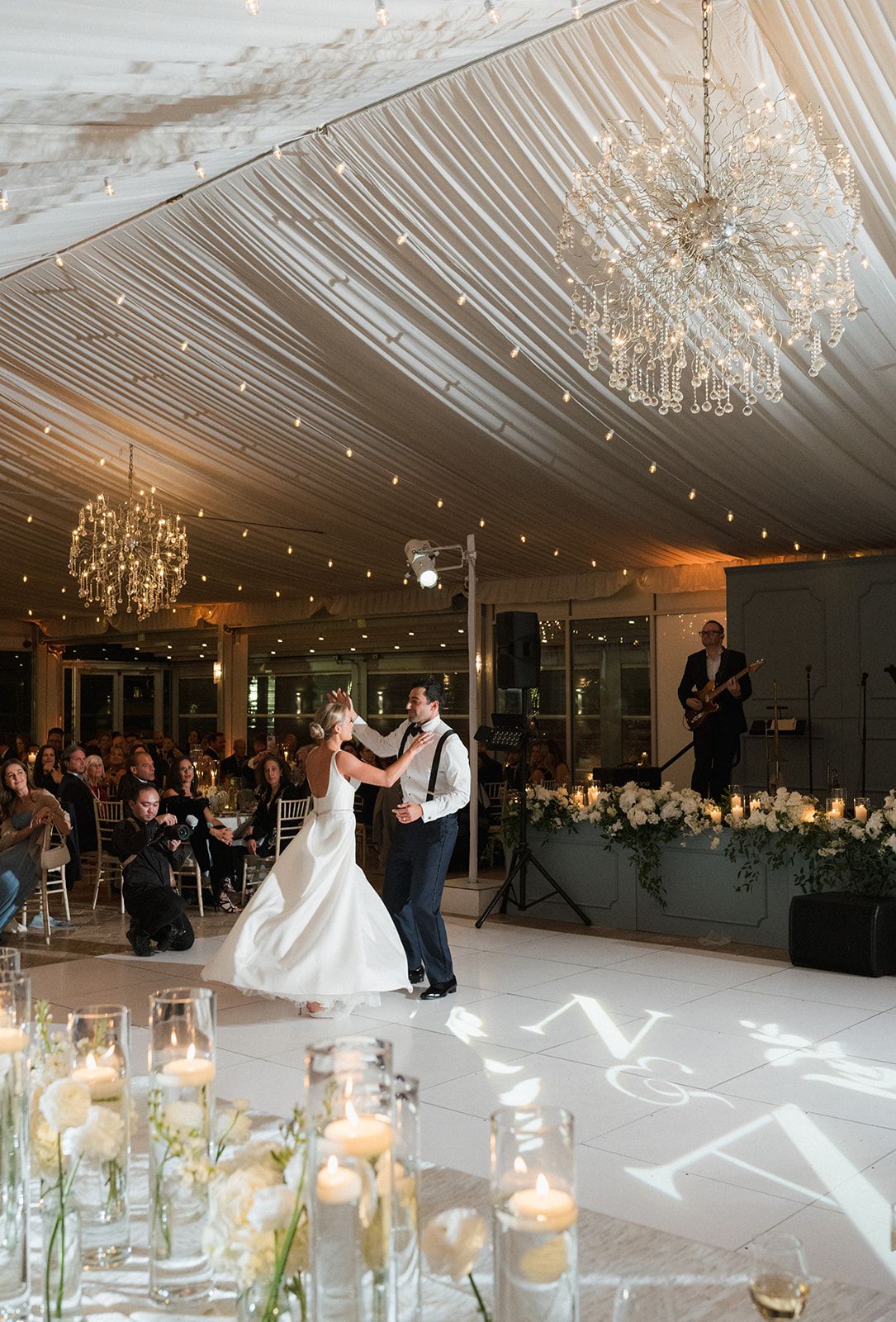 Bride and groom share first dance under crystal chandeliers with monogram floor projection at reception.