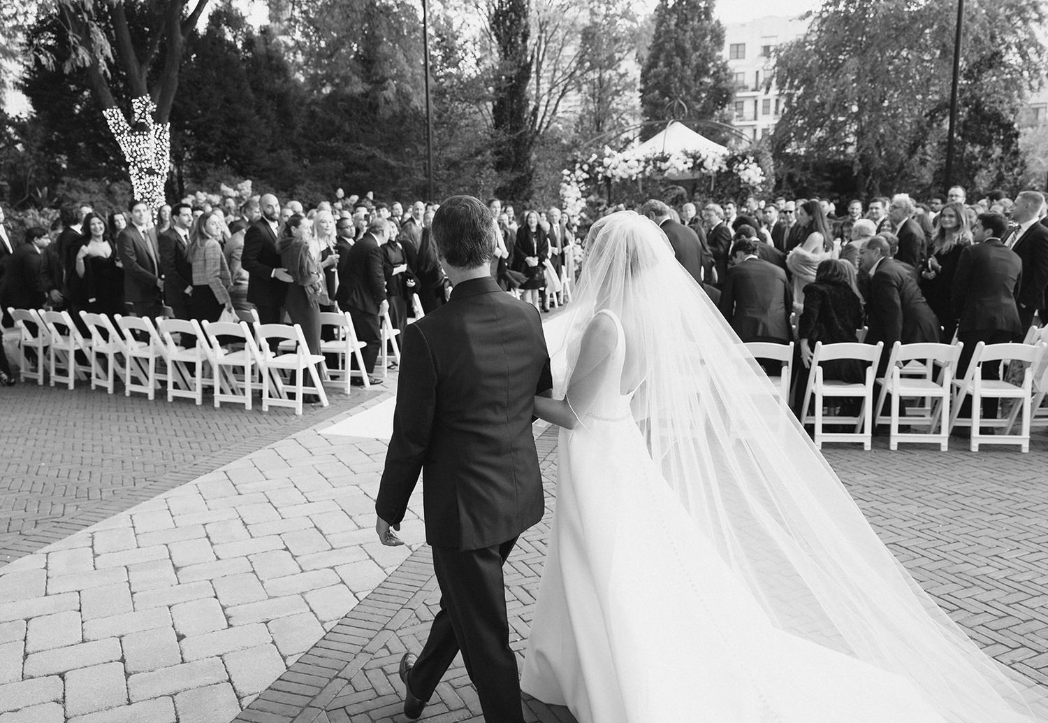 Black and white photo of bride and groom walking hand in hand toward outdoor wedding ceremony chairs.