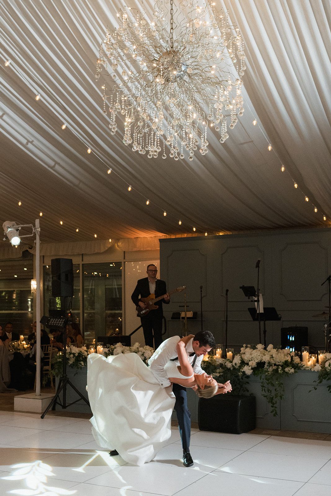 Groom dips bride during first dance under a crystal chandelier in an elegant white draped reception venue.
