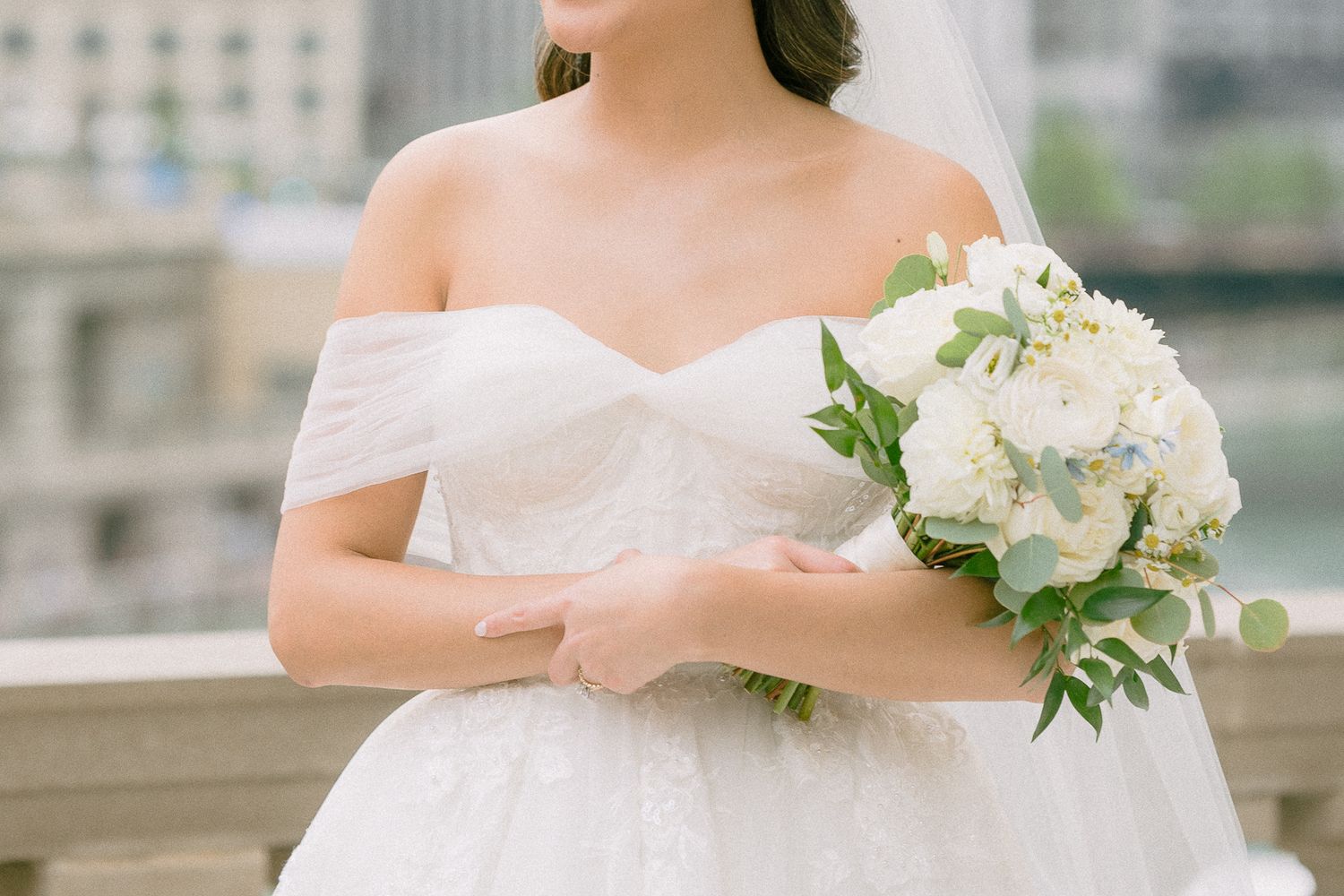 Bride in off-shoulder white ballgown holding lush white and green bouquet on outdoor balcony.