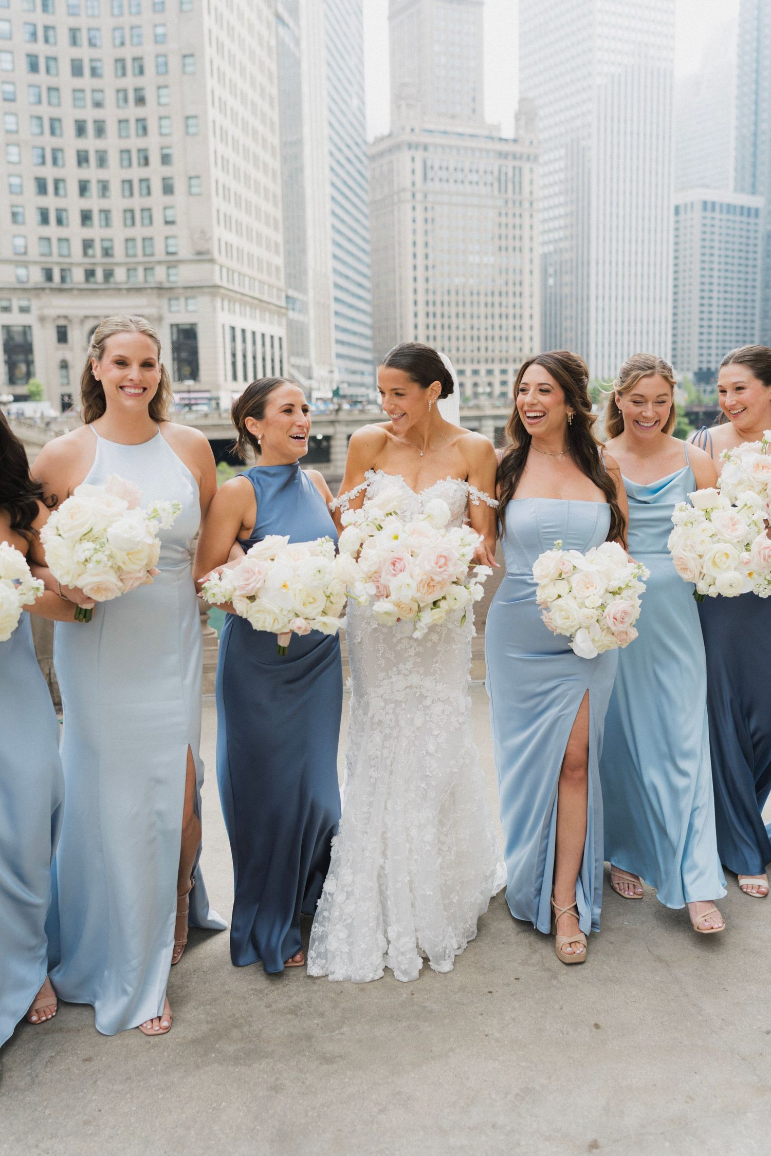 Bride in white lace gown walks with bridesmaids in blue satin dresses holding white bouquets with city skyline backdrop.