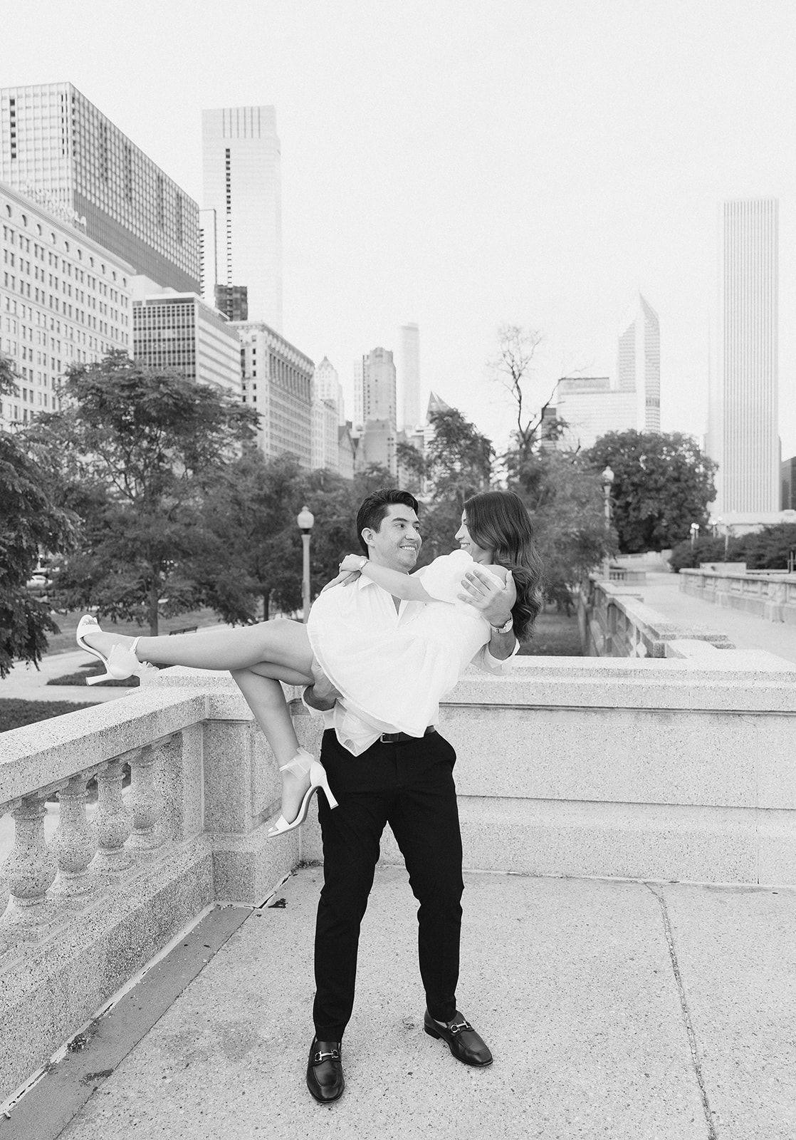 Groom lifting bride in white dress with Chicago skyline backdrop in black and white engagement photo.