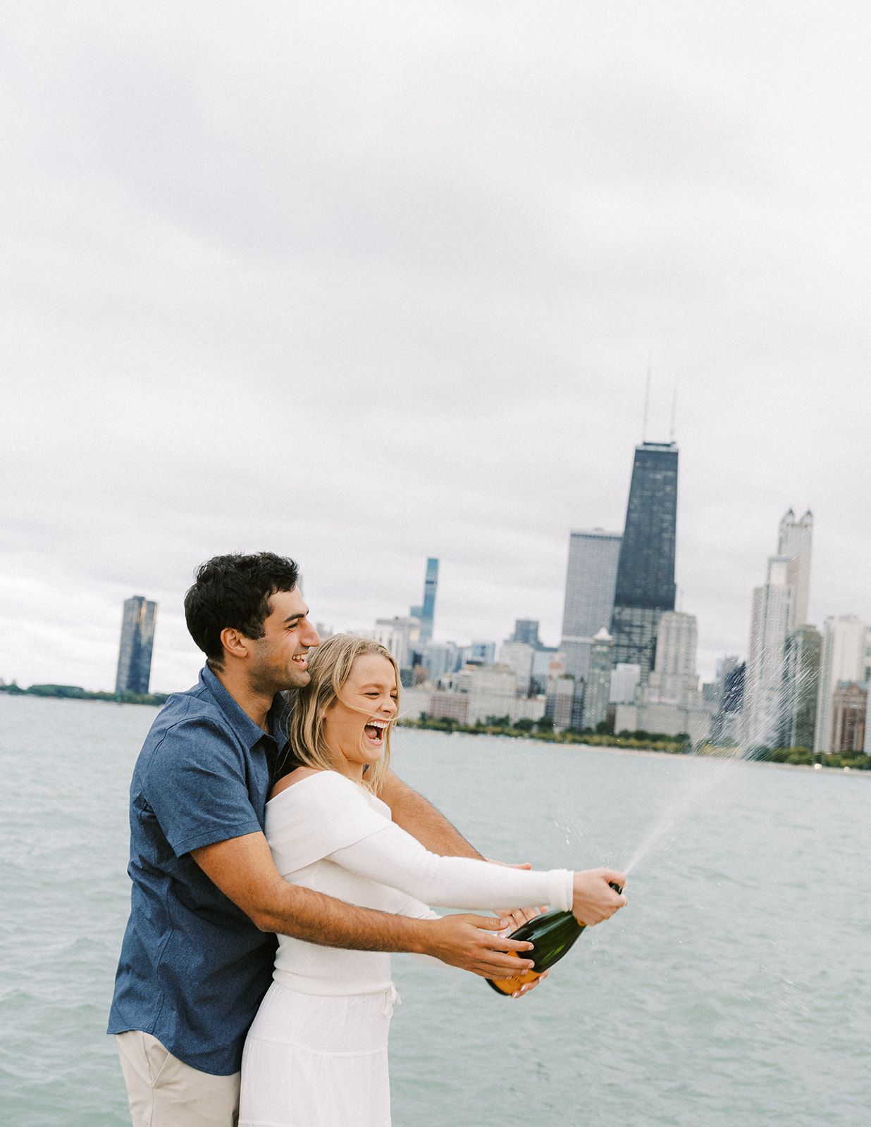 Couple celebrating engagement by popping champagne bottle on Chicago lakefront with city skyline.