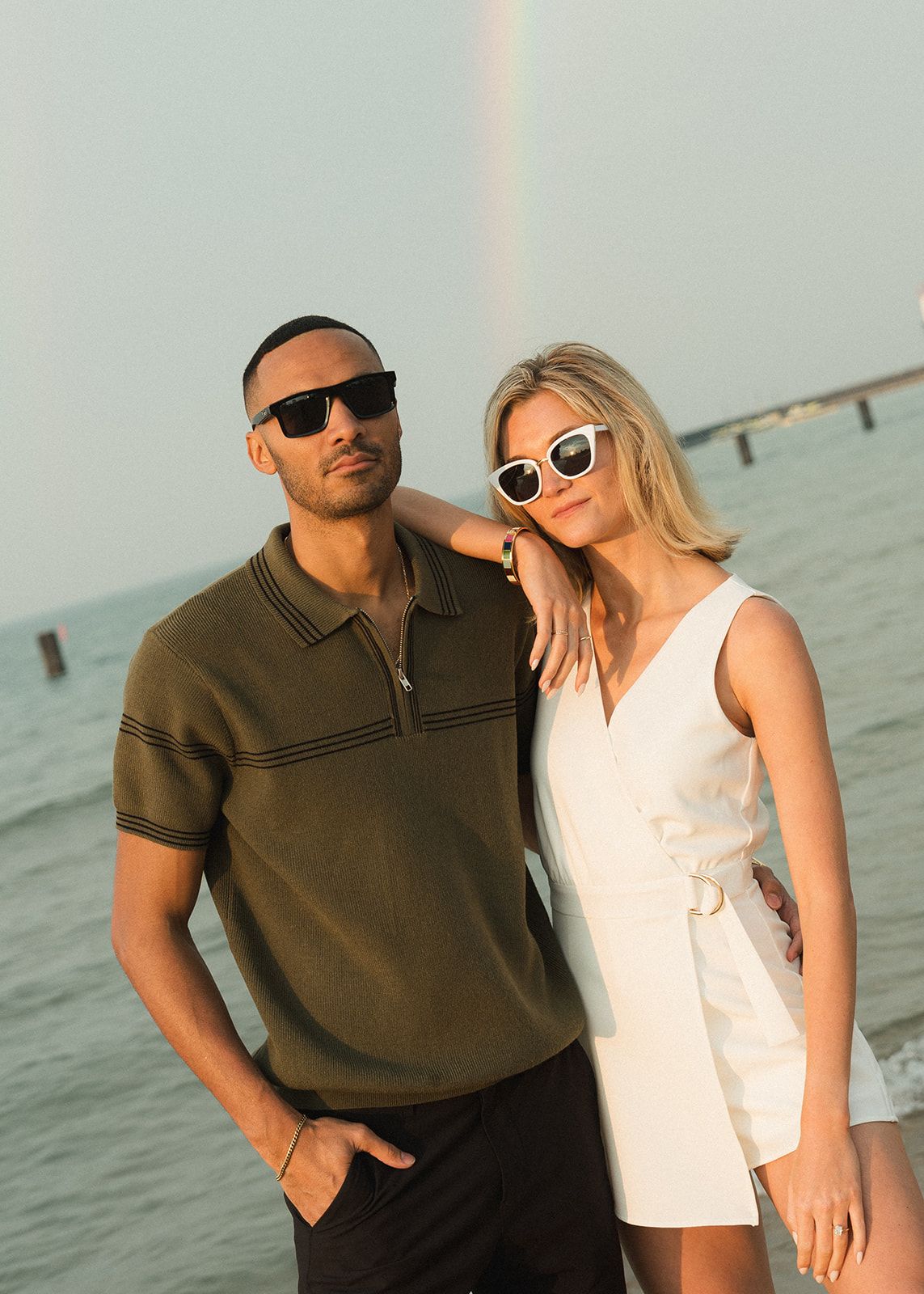 Stylish couple wearing sunglasses posing together on a beach with ocean backdrop at dusk.
