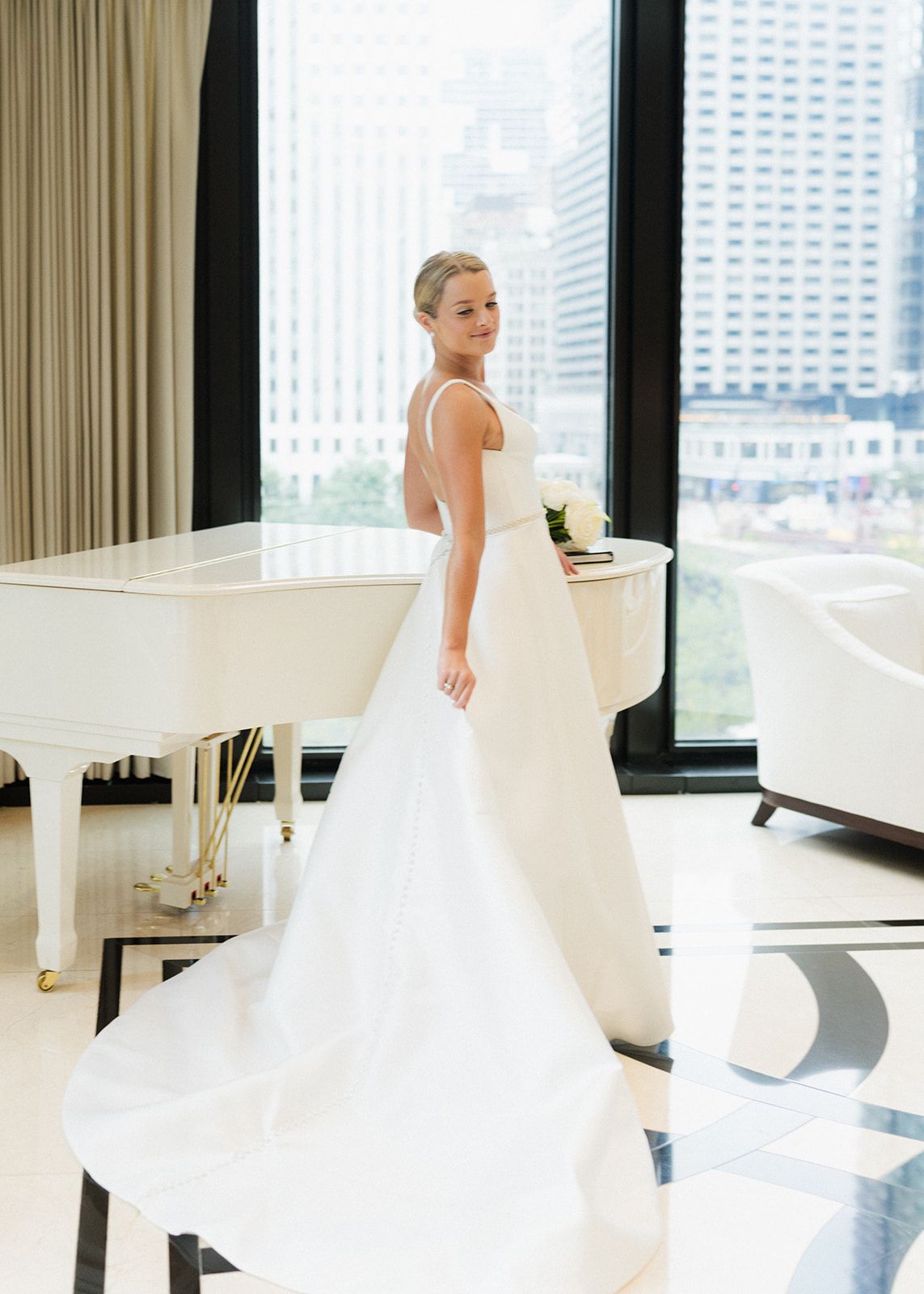 Bride in sleek white ball gown with open back stands near grand piano with city view.