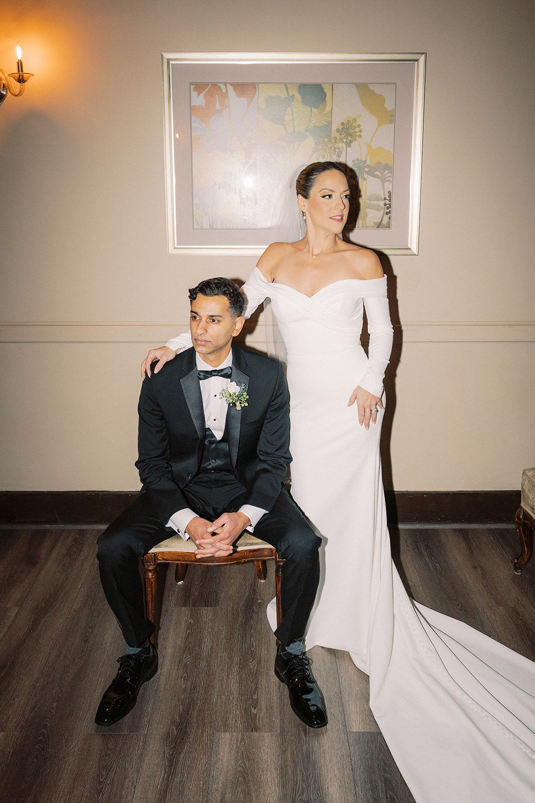 Bride in off-shoulder white gown stands beside seated groom in black tuxedo for wedding portrait.