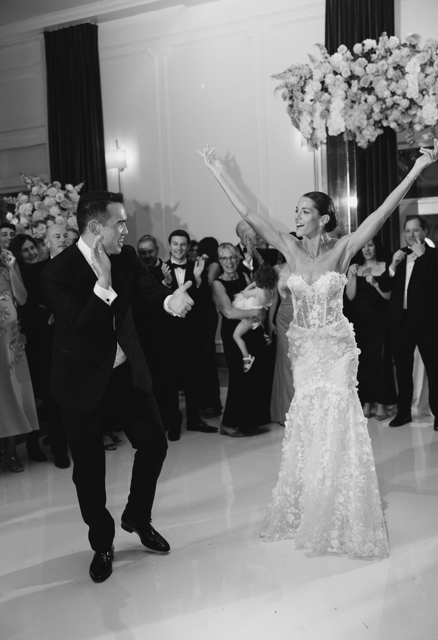 Joyful bride in floral gown raises arms as groom dances beside her at elegant black-tie wedding reception.