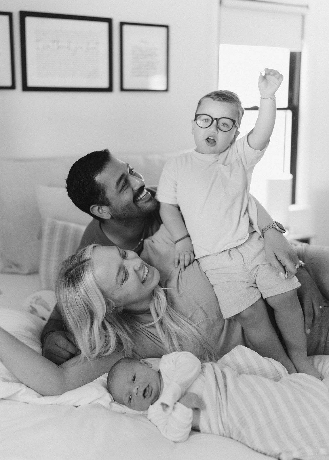 Black and white family photo of parents laughing with toddler in glasses and newborn baby on a bed.