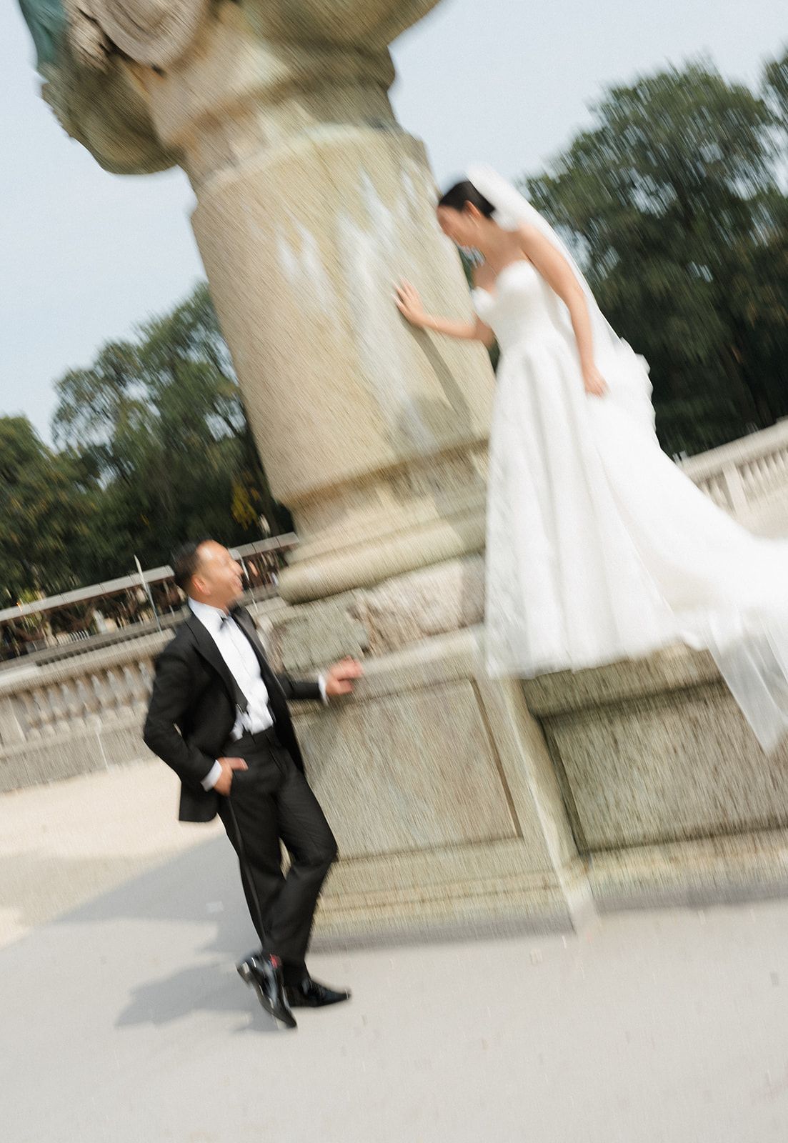 Motion-blurred wedding photo of bride in white gown standing on fountain ledge reaching toward groom outdoors.