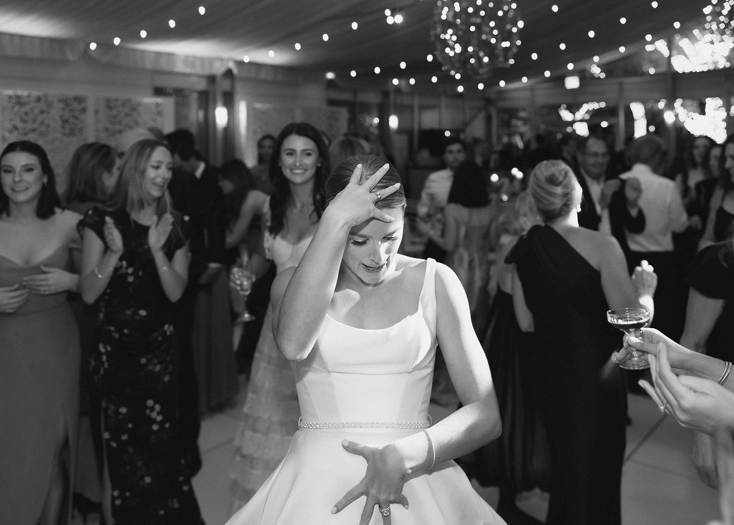 Black and white photo of a bride dancing joyfully at her wedding reception surrounded by smiling guests.