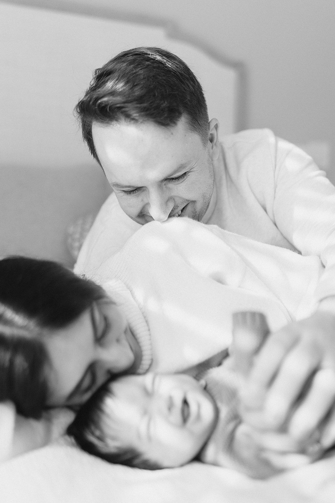 Black and white photo of smiling parents lovingly gazing at their laughing newborn baby on a bed.