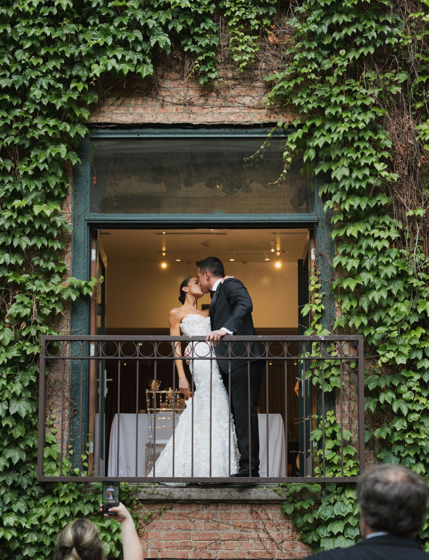 Bride and groom share a kiss on an ivy-covered balcony during their outdoor wedding ceremony.