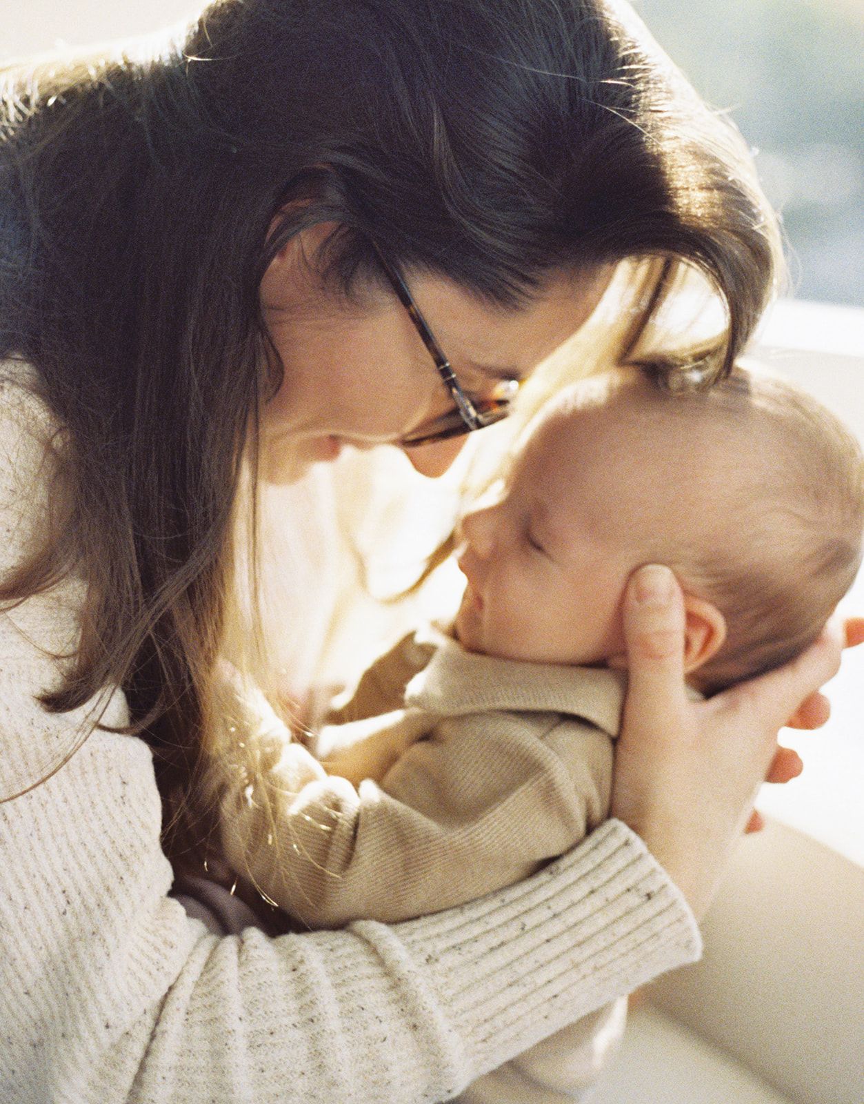 Tender moment of dark-haired mother with glasses gently cradling sleeping newborn baby close.