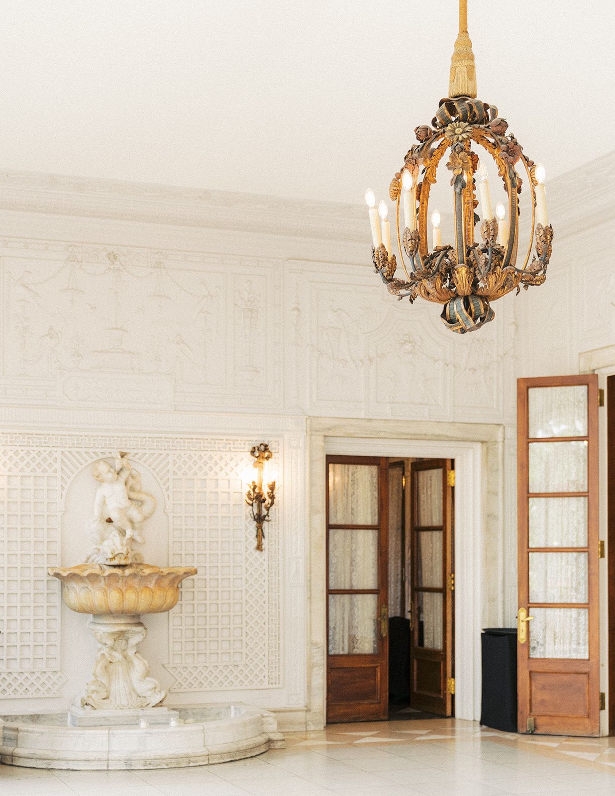 Elegant white marble foyer with ornate gold chandelier, classical fountain sculpture, and wooden doors.