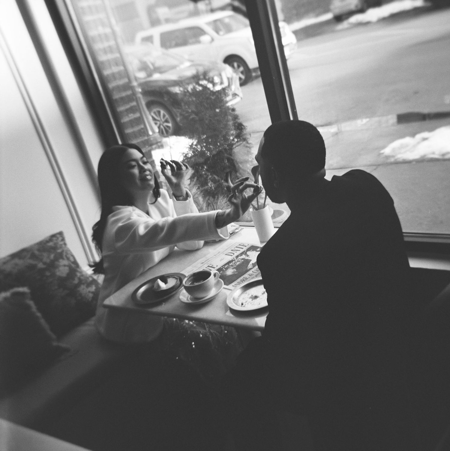 Black and white photo of a couple sharing coffee and laughter at a cafe table by a window.