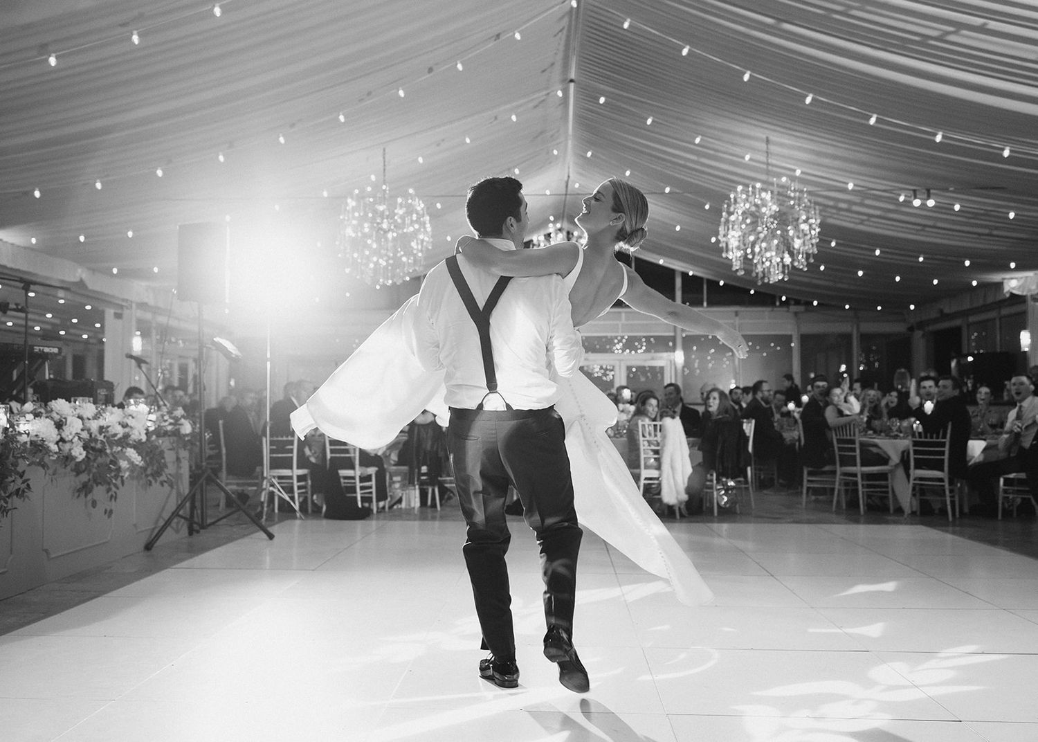 Black and white photo of groom lifting bride during their first dance in a tent reception venue.