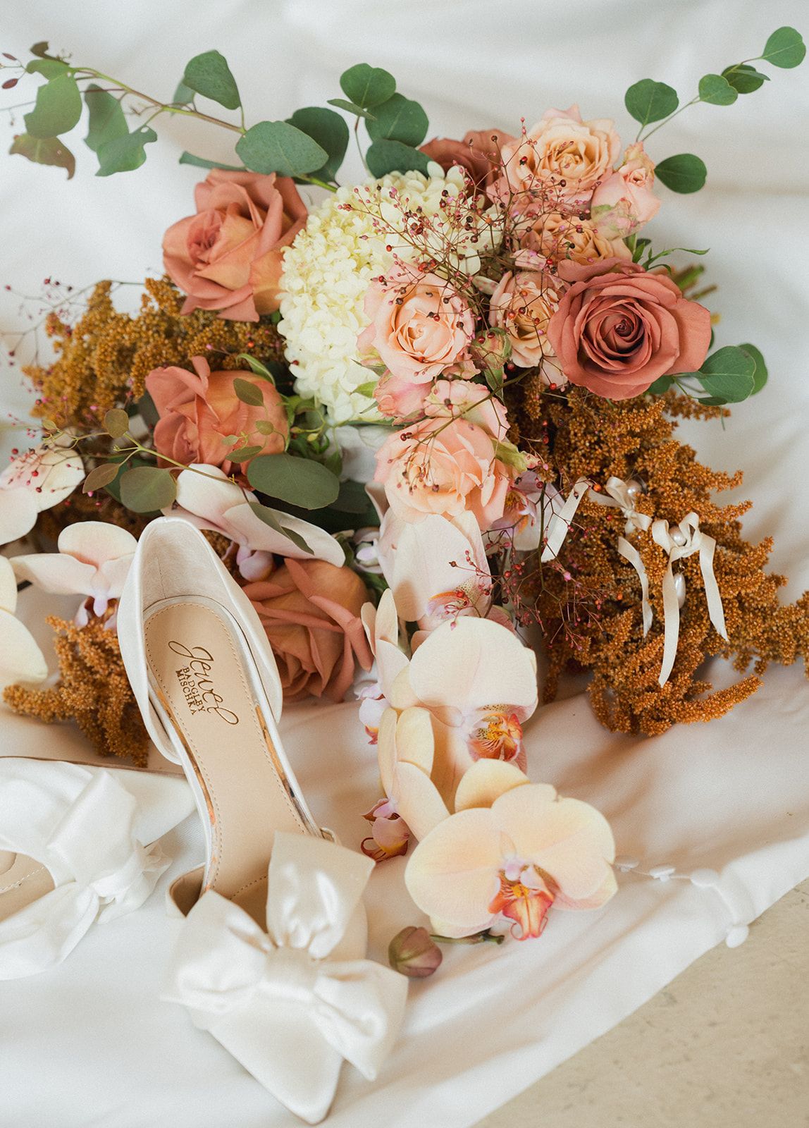 Bridal flat lay featuring ivory heels, peach and dusty rose bouquet with eucalyptus, orchids, and golden wildflowers on white fabric.
