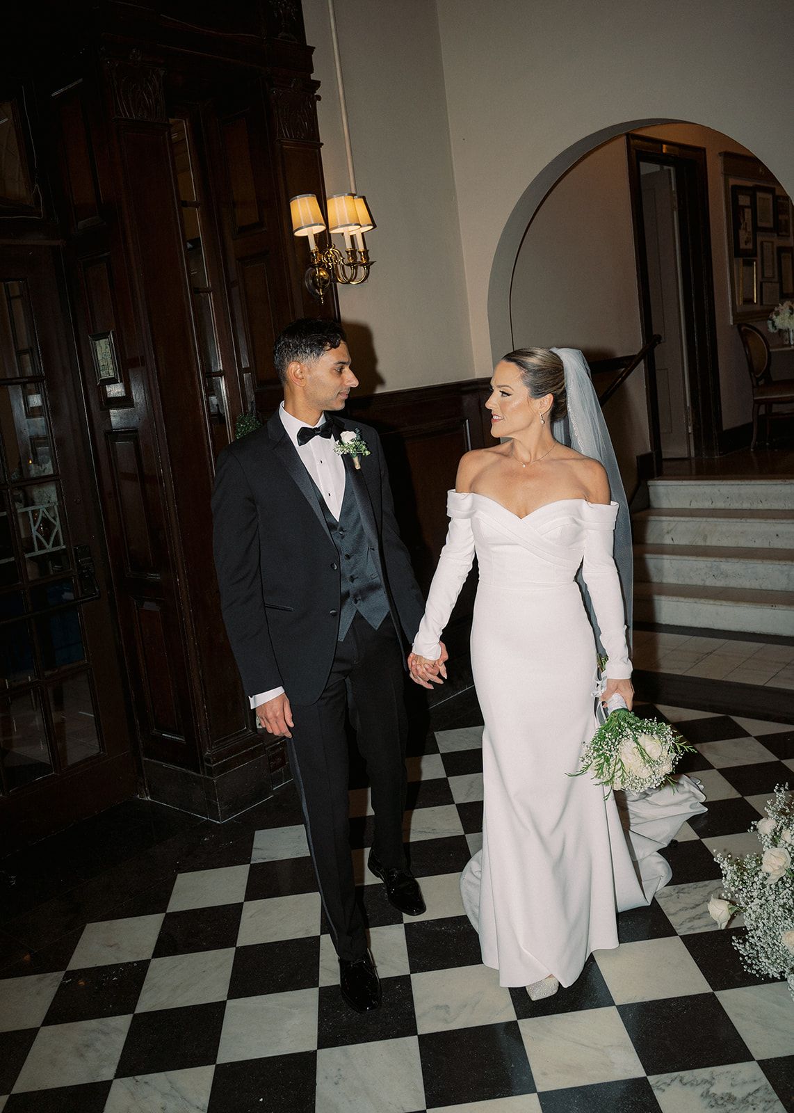 Bride in off-shoulder white gown and groom in dark tuxedo hold hands on black and white checkered marble floor.