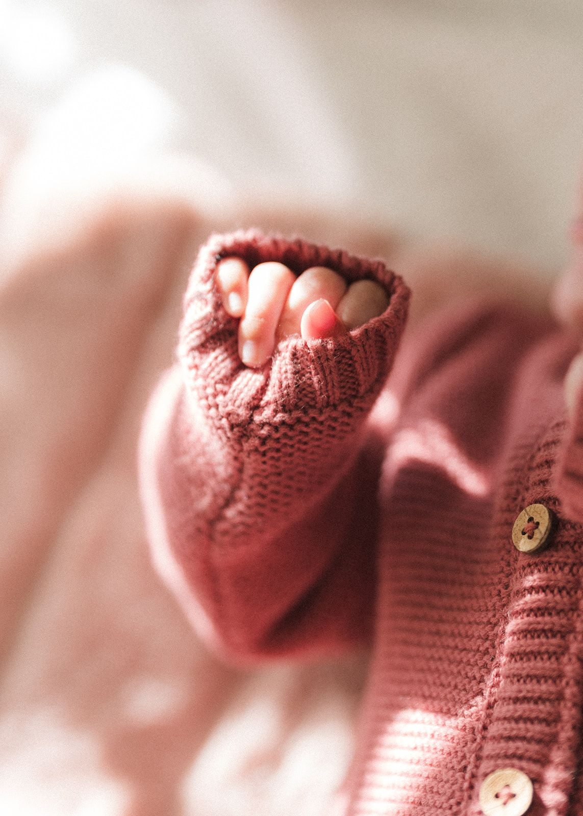 Close-up of a newborn baby's tiny fingers peeking out of a pink knit cardigan sleeve with wooden buttons.