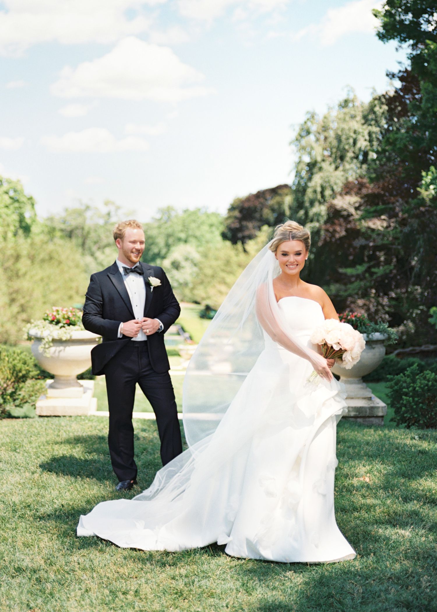 Bride in strapless white gown with veil holds bouquet as groom in black tuxedo stands behind her in lush garden.