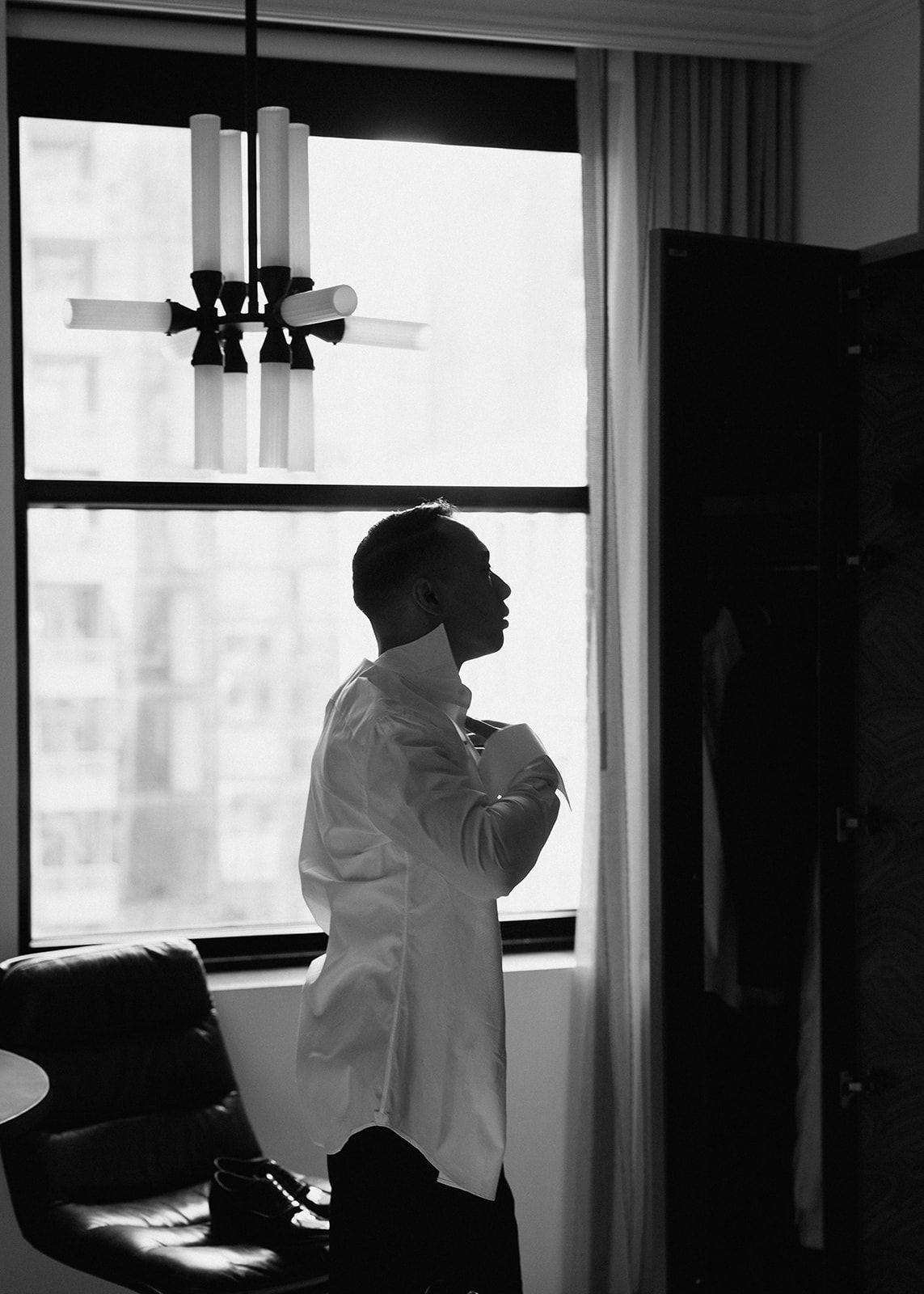 Black and white silhouette of groom getting dressed by a window before his wedding ceremony.