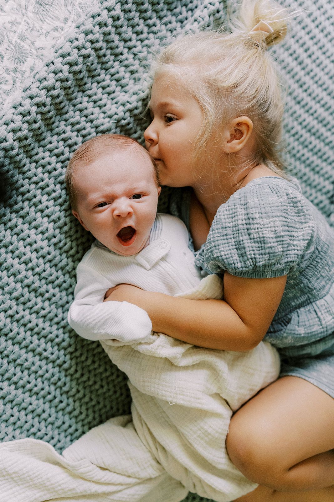 Toddler girl with blonde hair kissing yawning newborn baby sibling on teal knit blanket.