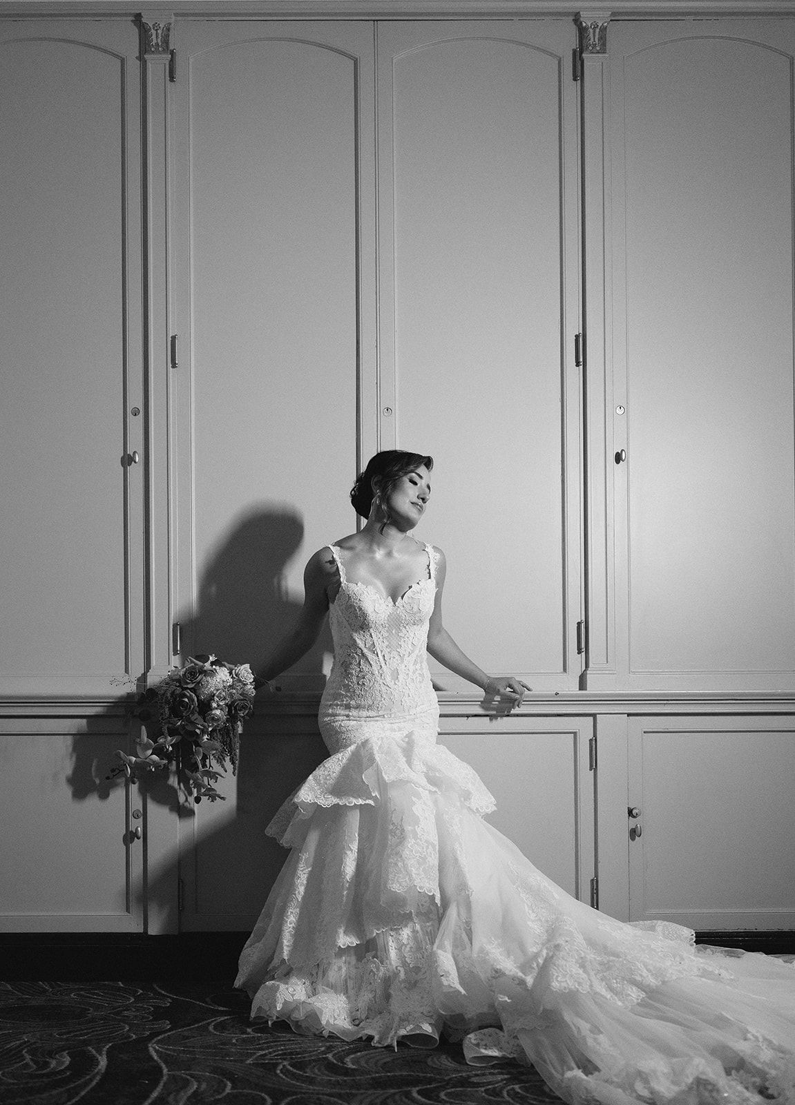Bride in a mermaid lace wedding gown holding a bouquet, posing elegantly against ornate white doors in B&W.