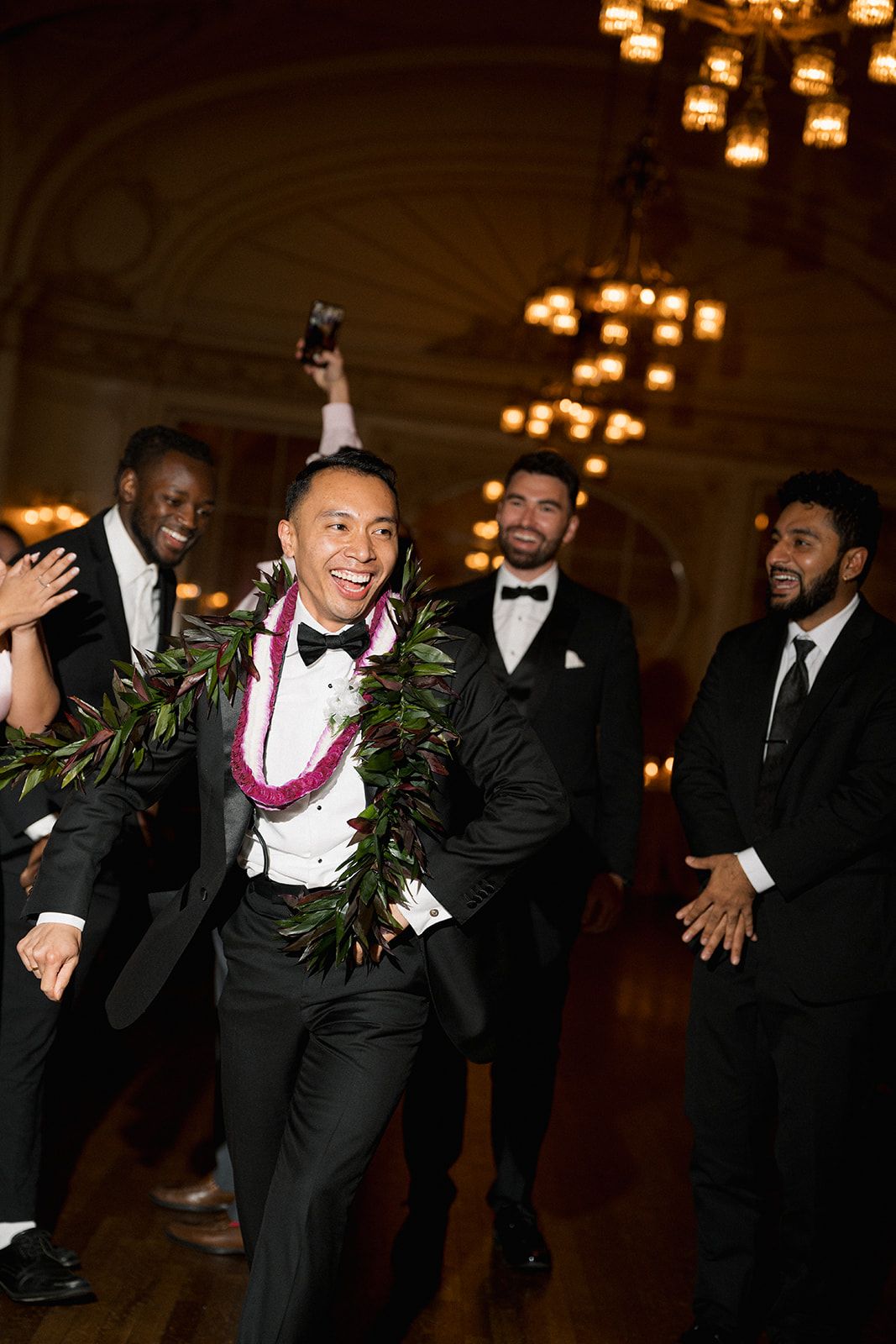 Groom wearing floral lei celebrates with groomsmen in black tuxedos in warmly lit ballroom reception venue.