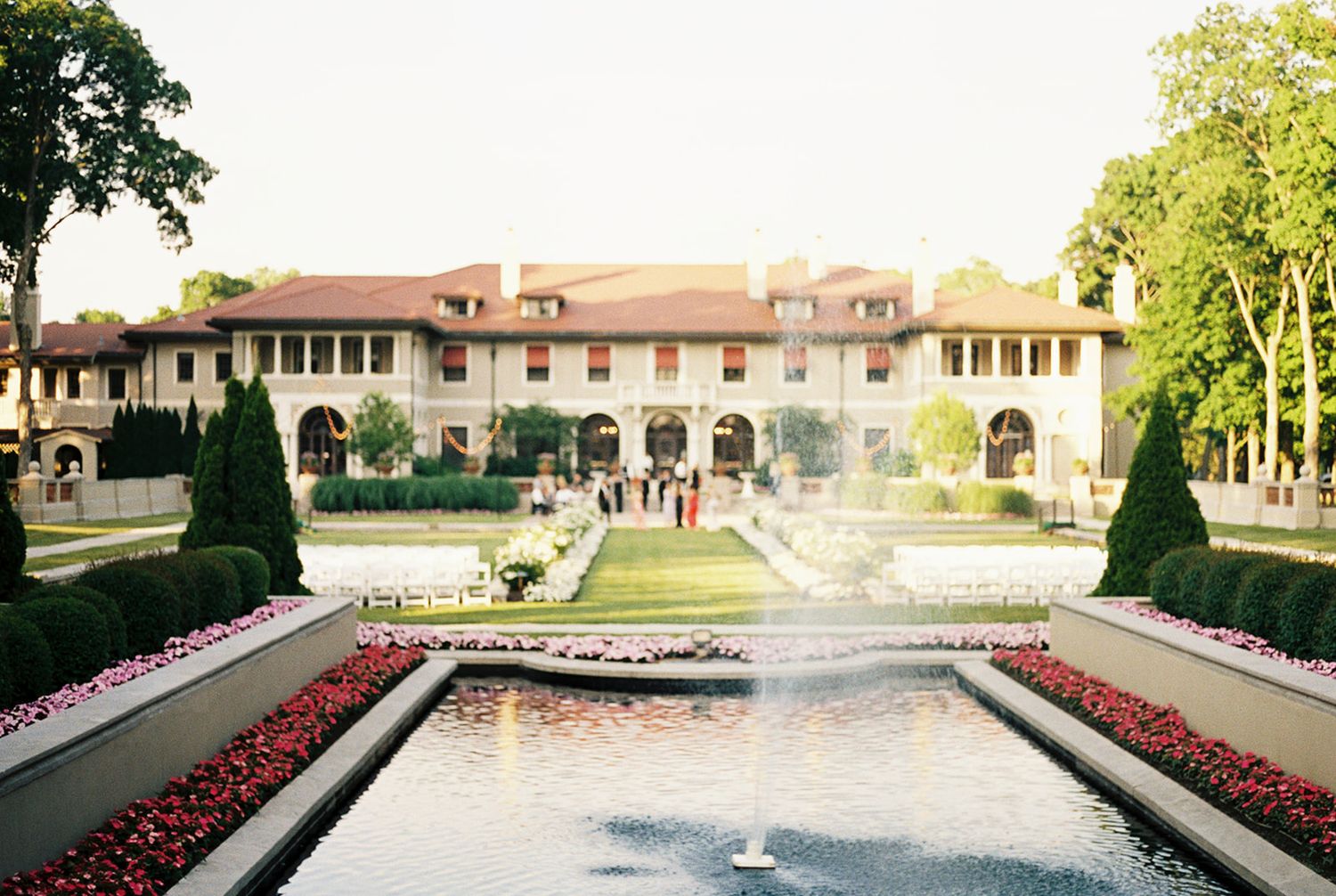 Elegant mansion wedding venue with fountain, manicured hedges, and red flower borders on a sunny day.