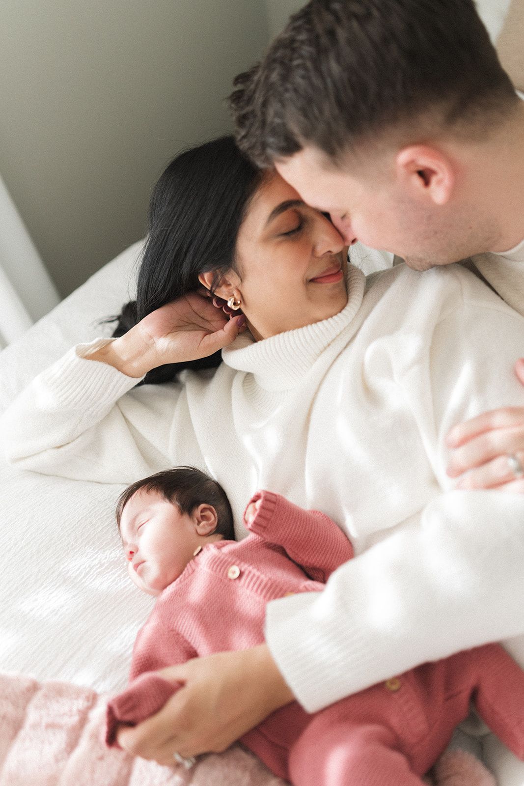 A couple in white sweaters cuddles on a bed with their sleeping newborn baby dressed in a pink knit outfit.